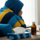 Person wrapped in a blanket sitting on a couch with tissues, cough medicine, and a glass of water while feeling unwell.