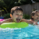 **Alt Text:** Children laughing and splashing together in a backyard swimming pool on a sunny summer day.