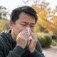 Asian man experiencing fall allergy symptoms and using a tissue outdoors during ragweed season.