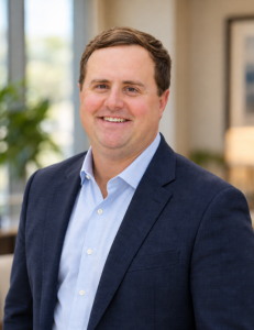 Drew Smith smiling in a navy blazer and light blue dress shirt in a professional office setting.