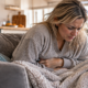 Woman with stomach cramps resting on a couch with water, soup, crackers, and an electrolyte drink nearby while a child relaxes in the background.